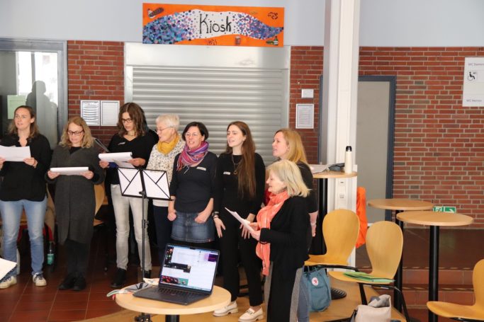 Eine Gruppe von Frauen singt zusammen in einem Raum mit Tischen und Stühlen.