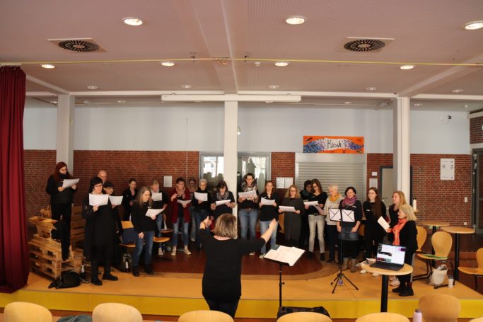 Ein Chor singt zusammen, geleitet von einer Dirigentin in einem Innenraum.